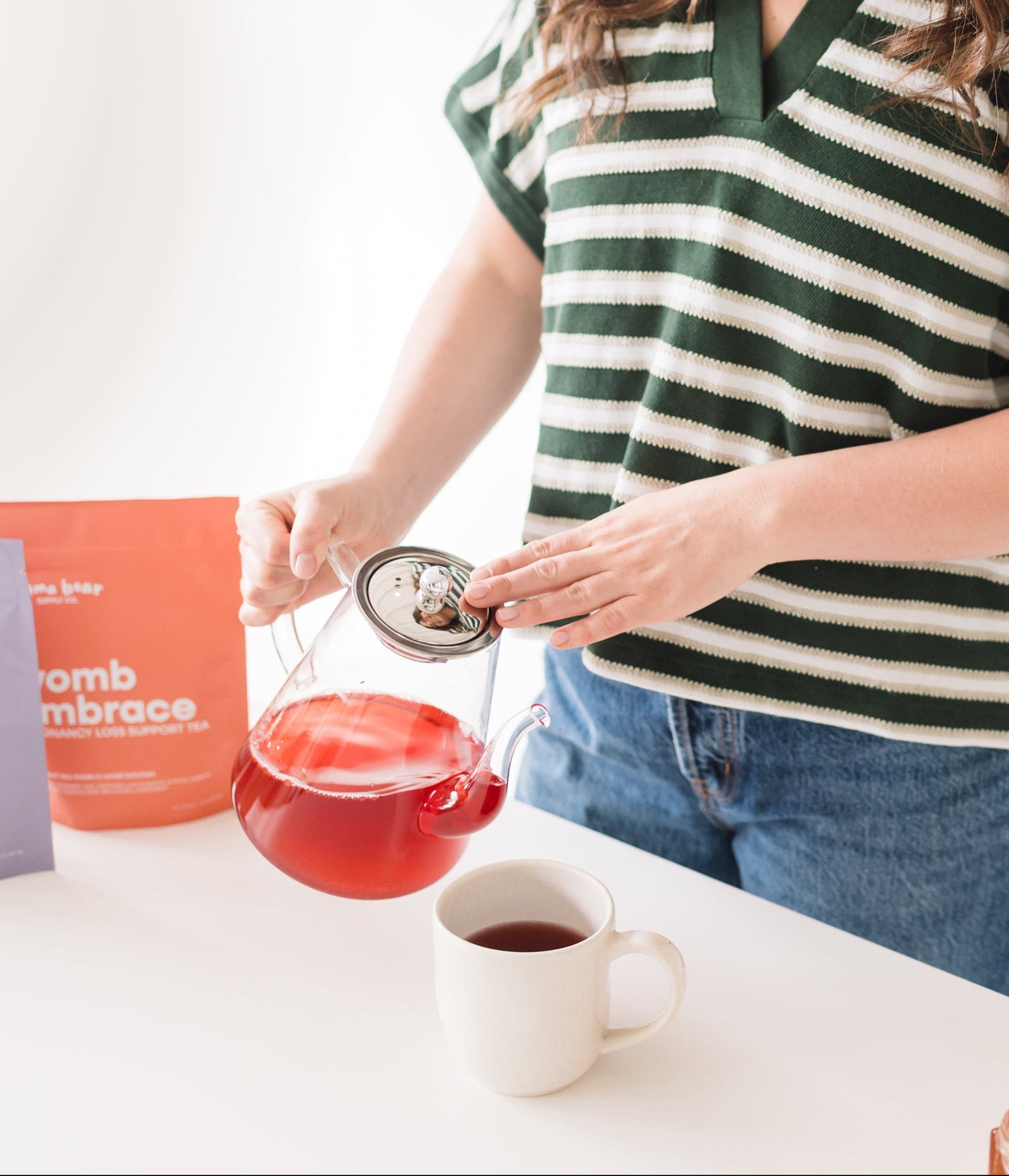 Woman pouring tea from a teapot into a cup on a white surface.
