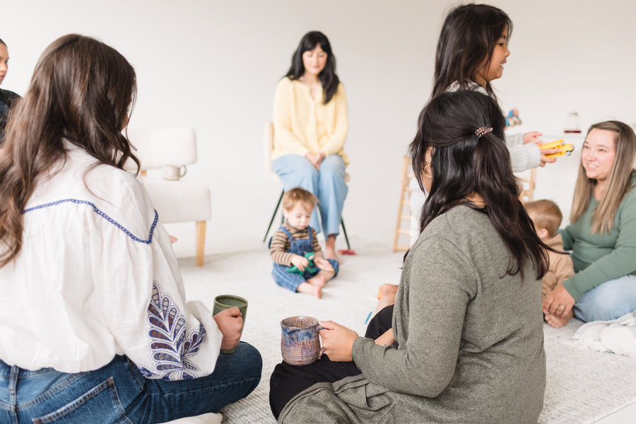 Group of women and children sitting on the floor in a circle, possibly in a home setting.